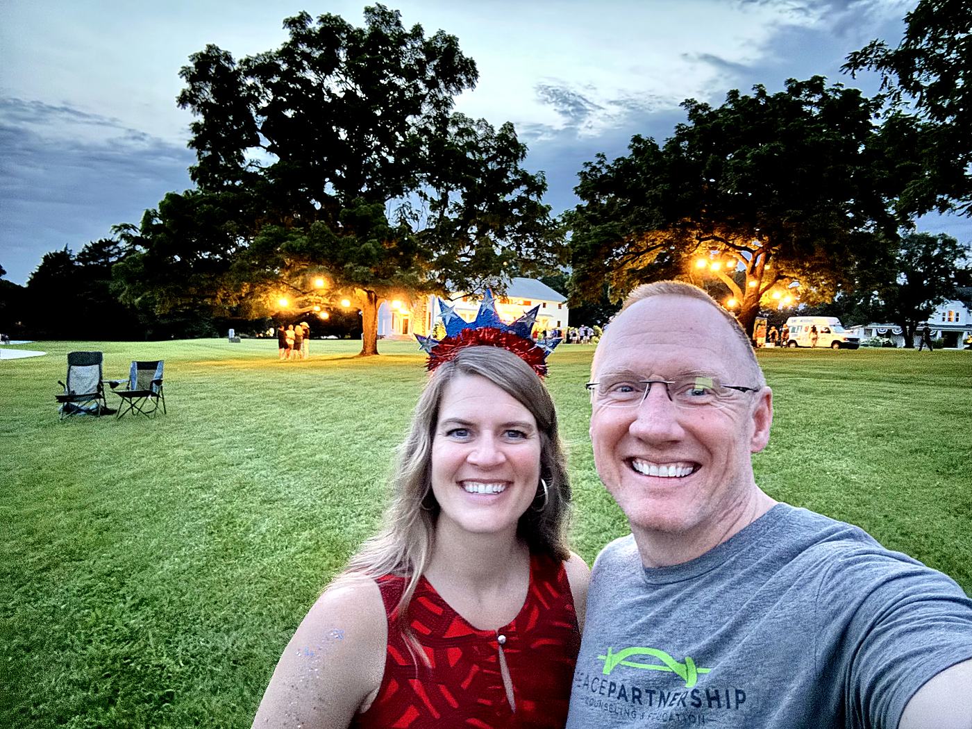 The two of us at a Fourth of July lawn party at dusk — star-spangled headband, lantern-lit trees, and a white farmhouse behind.