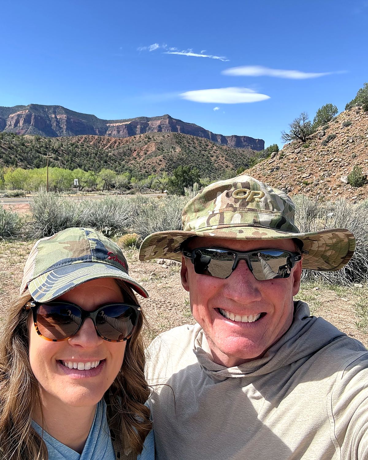 The two of us on a canyon road trip — matching sun hats and sunglasses, grinning, with red-rock cliffs and sagebrush behind.