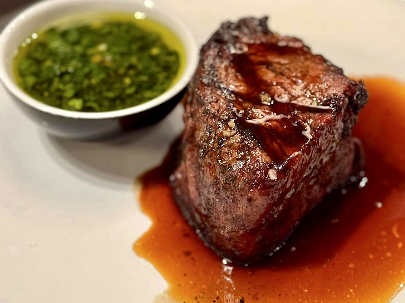 A dry-aged filet at Acre — deeply charred, resting in its own jus, with chimichurri alongside.