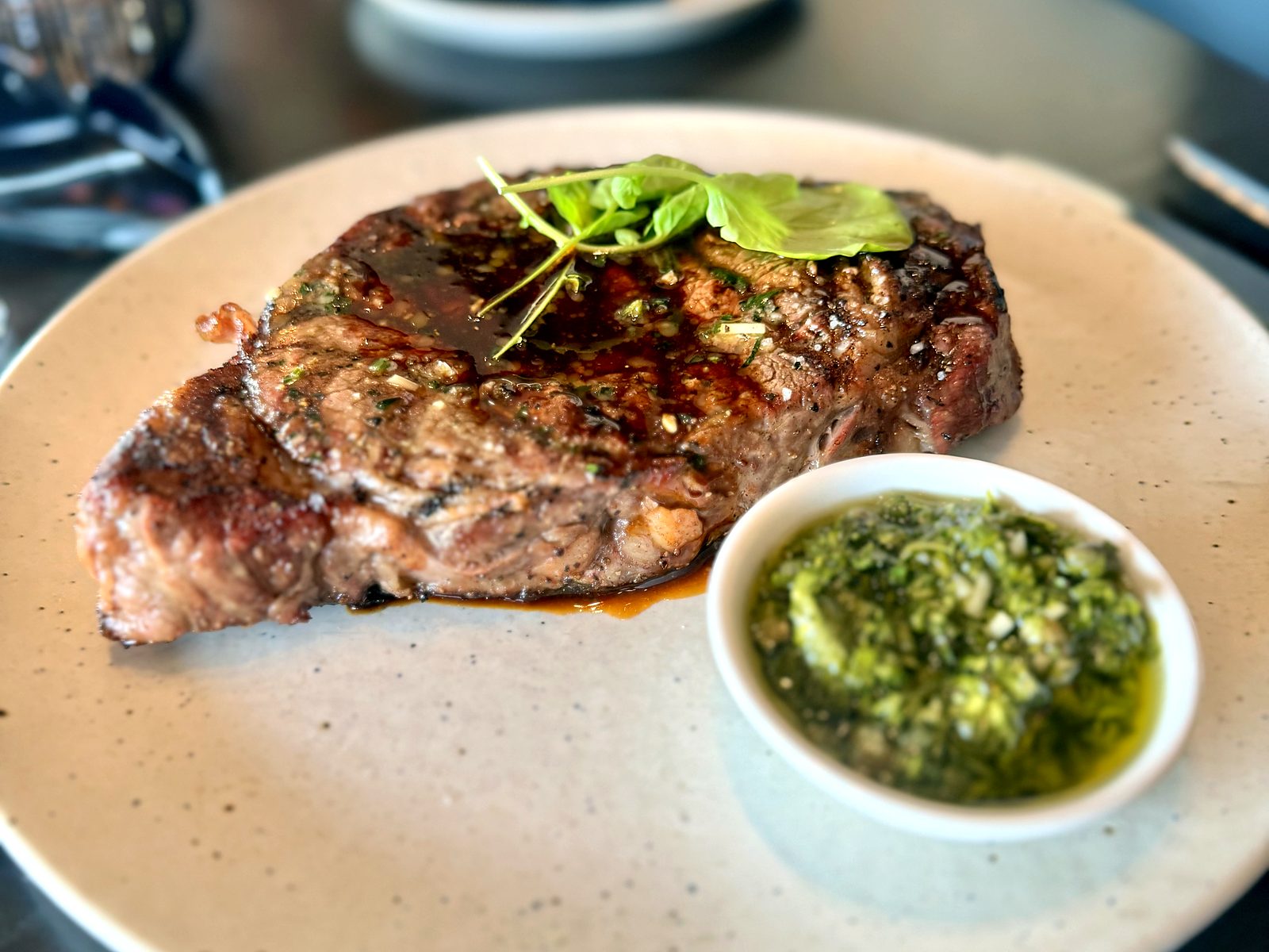 The dry-aged ribeye at Acre, basted and glistening under fresh basil, with a ramekin of chimichurri alongside.