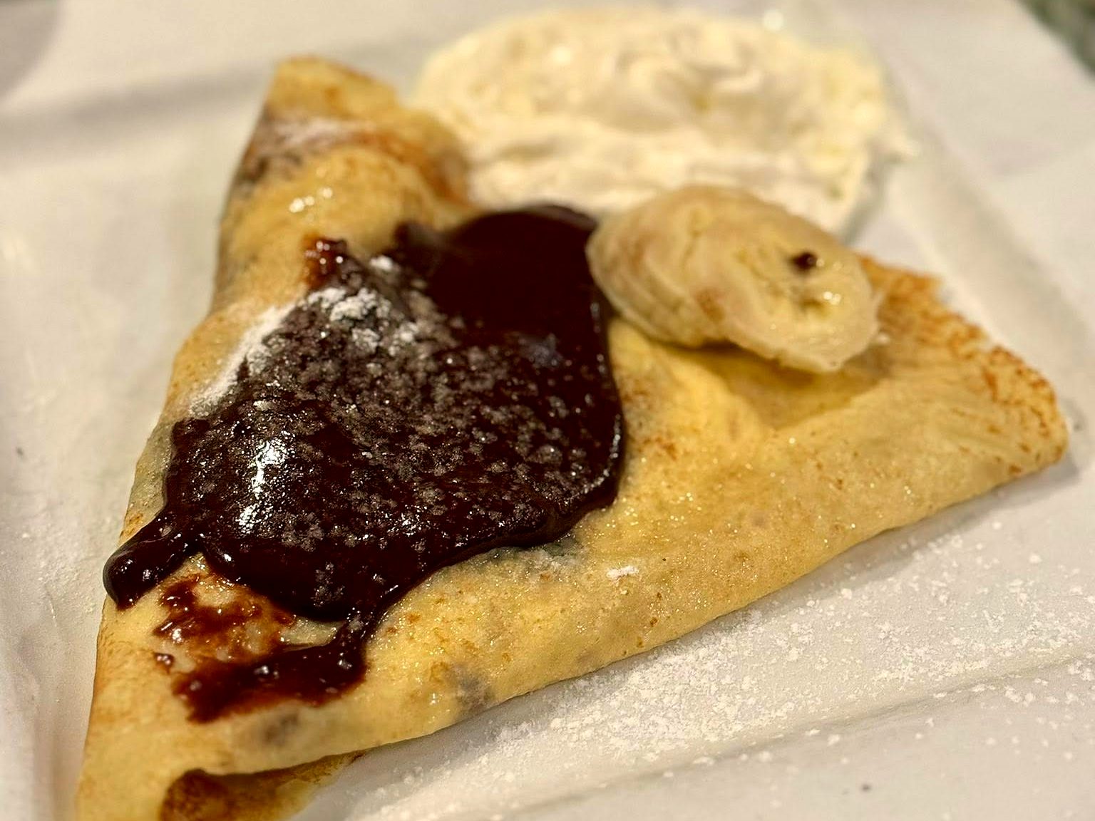 The chocolate crepe at Aixois — folded thin under a pour of dark chocolate, banana coins and whipped cream alongside, dusted with powdered sugar.