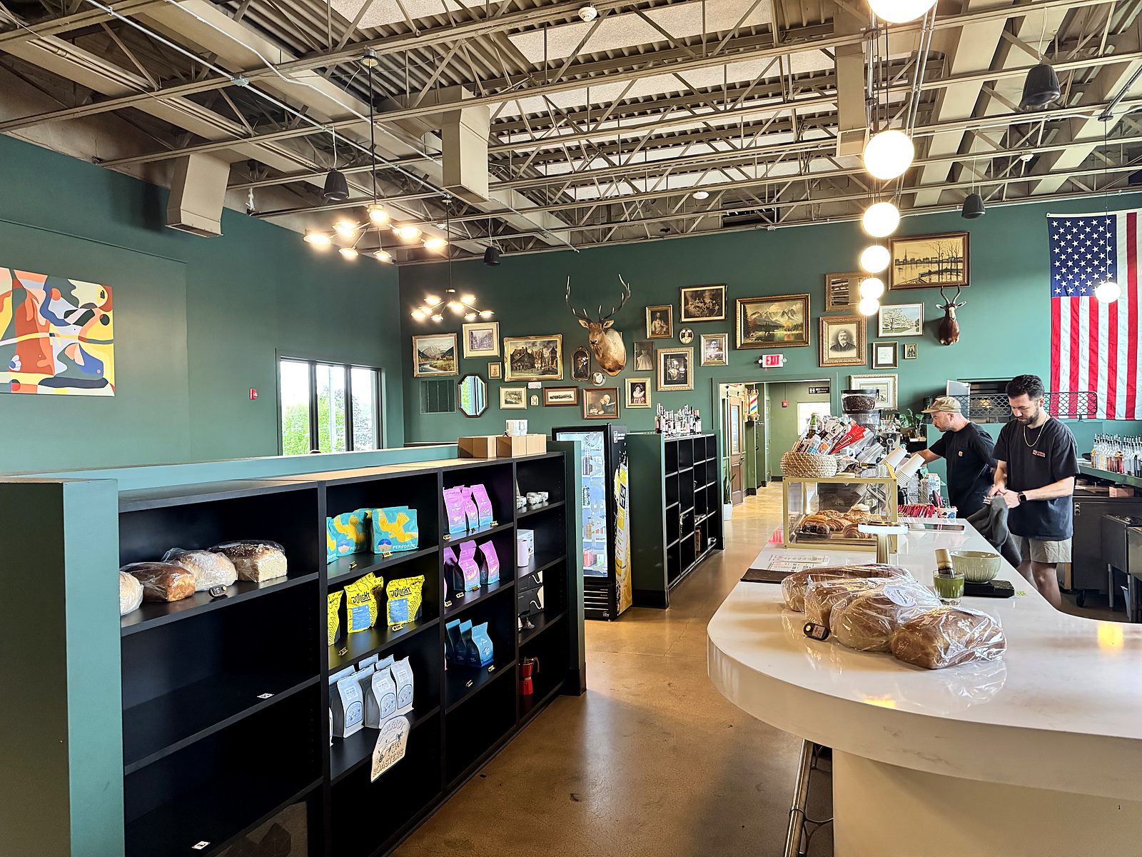 Inside Ase Coffee — a green-walled room hung with framed art and mounted antlers, retail shelves of coffee bags and bread, and two baristas working the curved white counter.