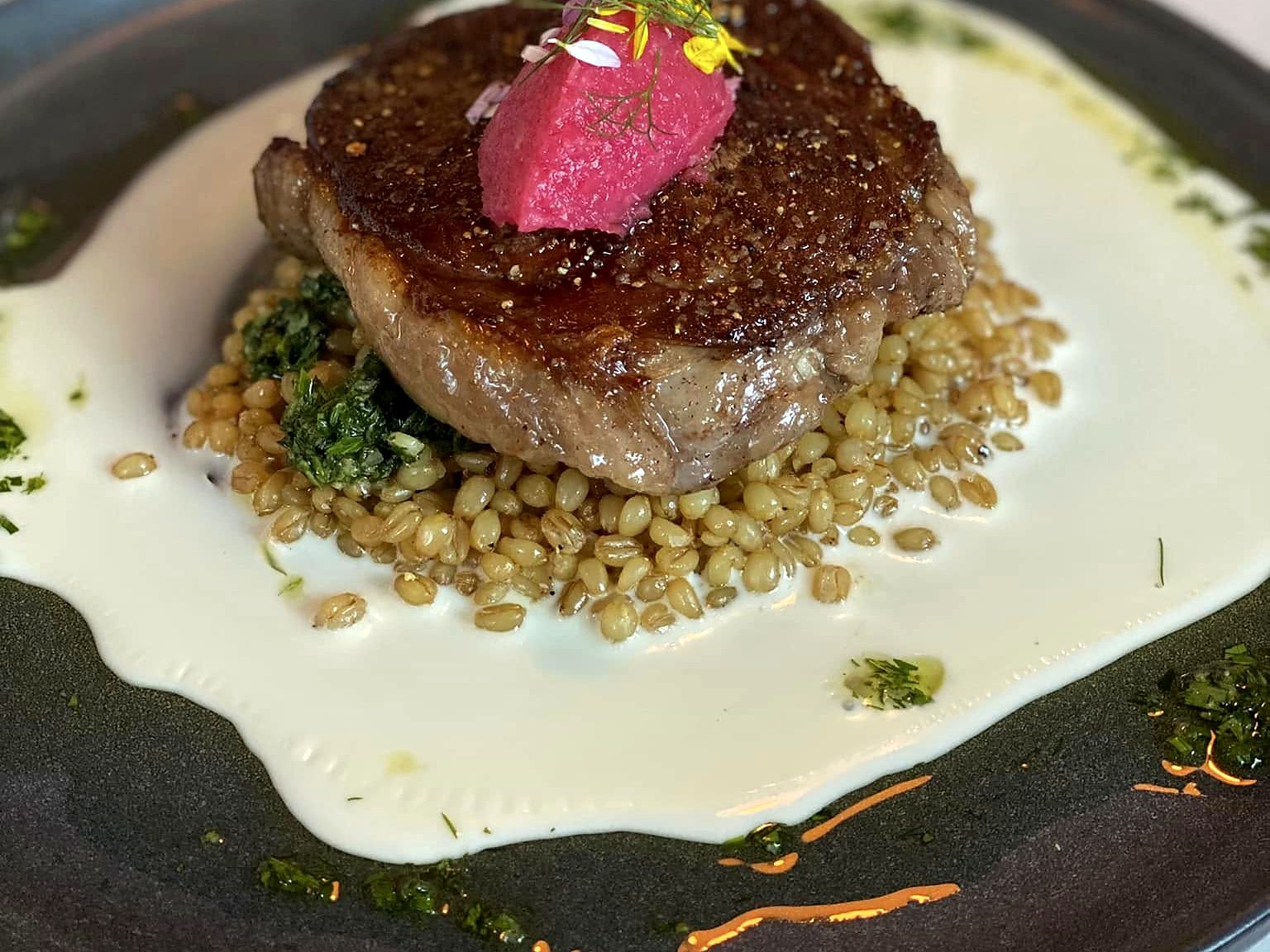 The bison ribeye at Bin 707 Foodbar — seared and peppered over wheat berries and salsa verde in a pool of cream, crowned with a beet-pink butter quenelle and flowers.