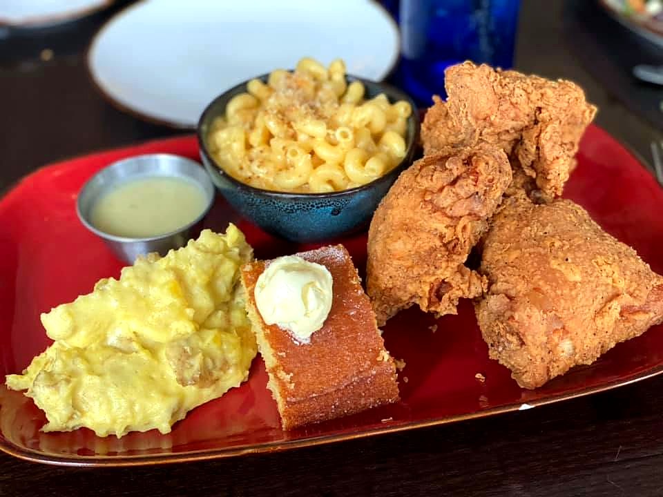 The fried chicken platter at The Brass Onion — craggy fried chicken with buttered cornbread, mac and cheese, mashed potatoes, and gravy on a red platter.