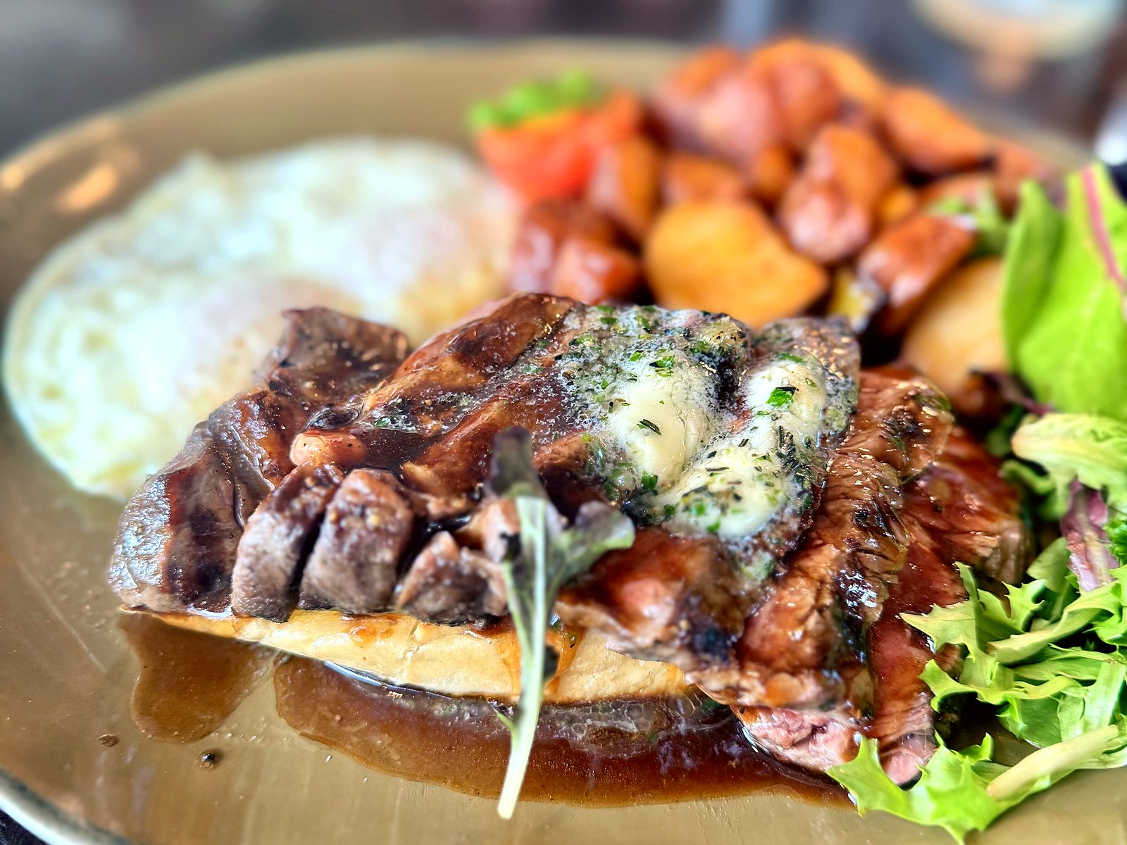 Steak and eggs at The Brass Onion — sliced steak over toast with melting herb butter and jus, a fried egg, breakfast potatoes, and dressed greens.