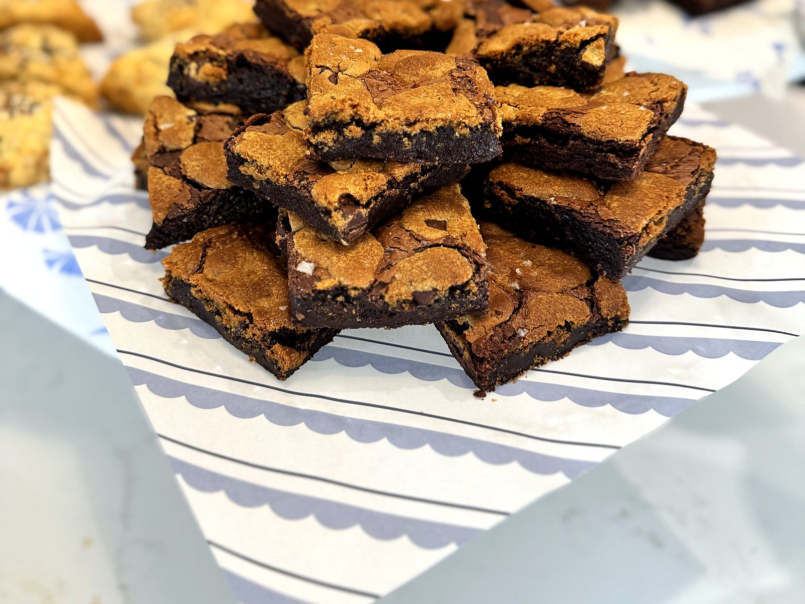 Cookie-brownie bars at Cornflower Baked Goods — chocolate chip cookie tops over fudge bases with flaky salt, piled on scalloped paper with the pastry counter behind.