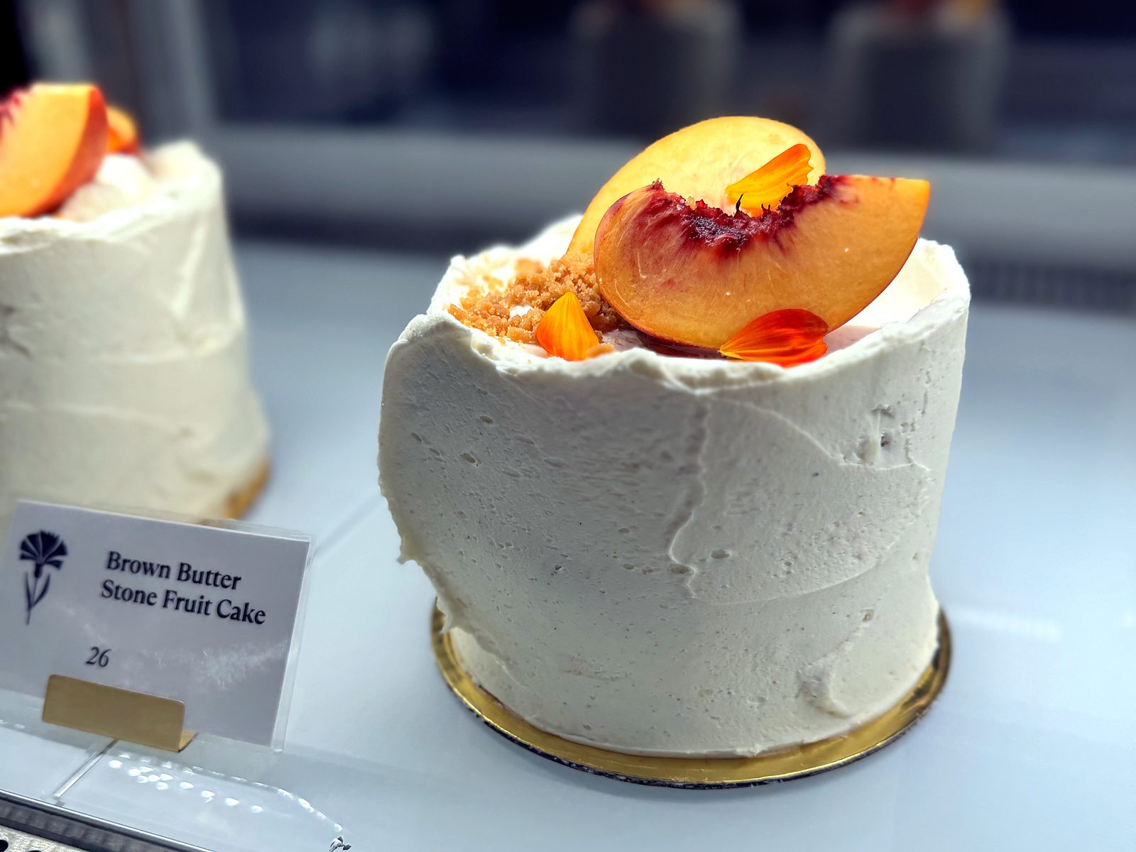 The brown butter stone fruit cake at Cornflower Baked Goods — a small white-frosted cake in the case, topped with fresh peach slices, crumble, and marigold petals.