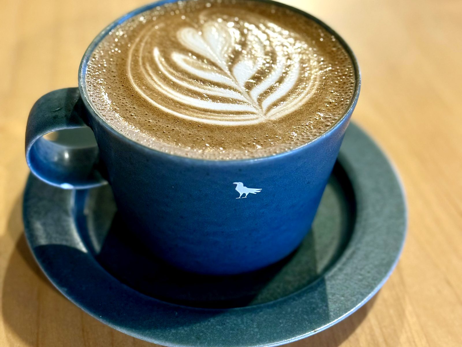 A latte at Corvus Coffee in a blue crow-logo cup, poured with a rosetta.