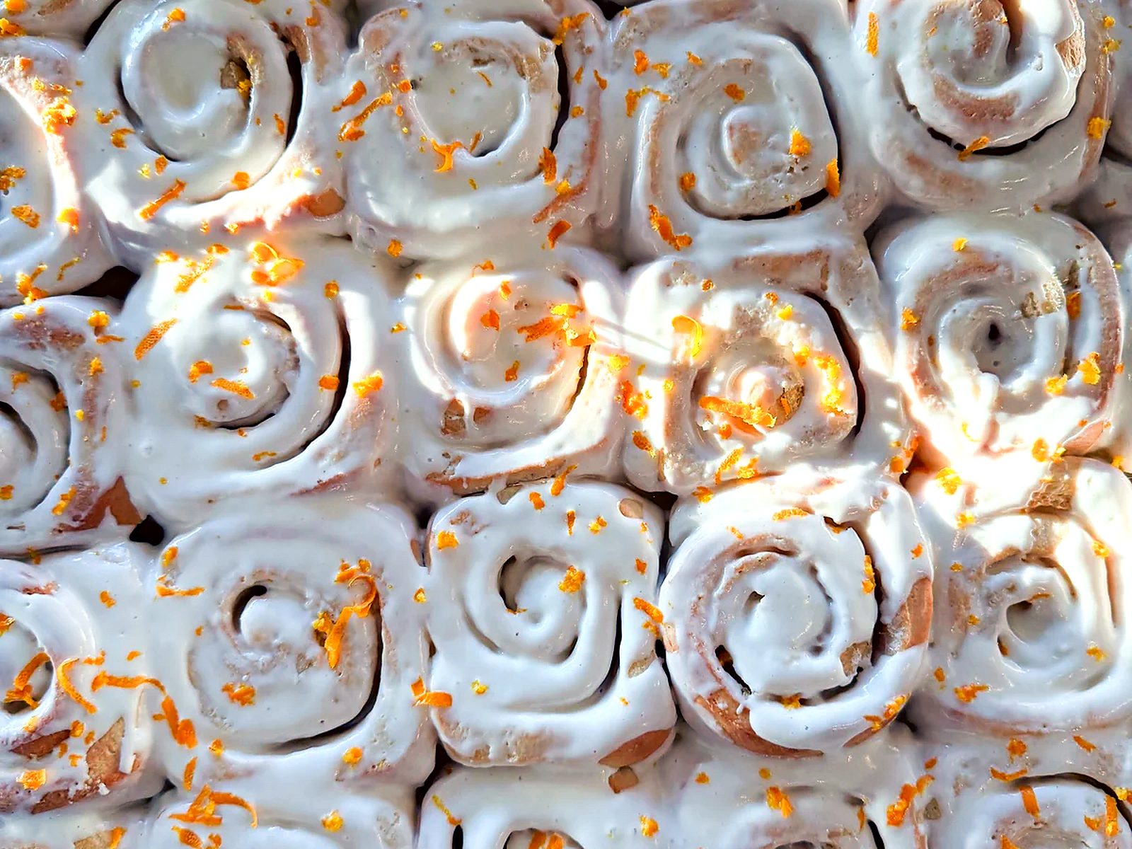A full tray of orange rolls at Dolce Bakery — iced spirals packed edge to edge, flecked with fresh orange zest.