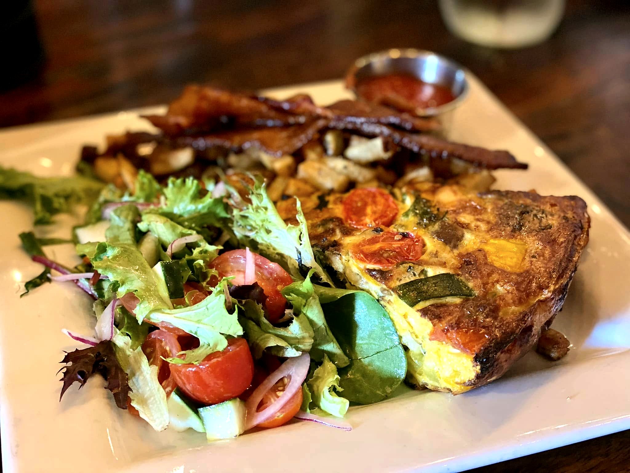 Brunch quiche at The Farmhouse — a browned vegetable wedge with dressed greens, candied bacon, and breakfast potatoes.