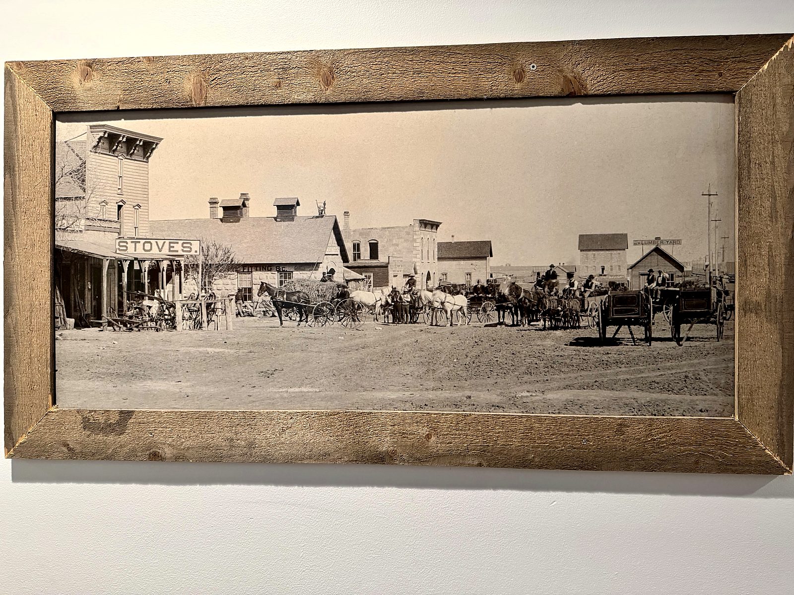 A framed historic photograph at Hickok&rsquo;s — a dirt main street lined with wagon teams and a storefront painted STOVES.