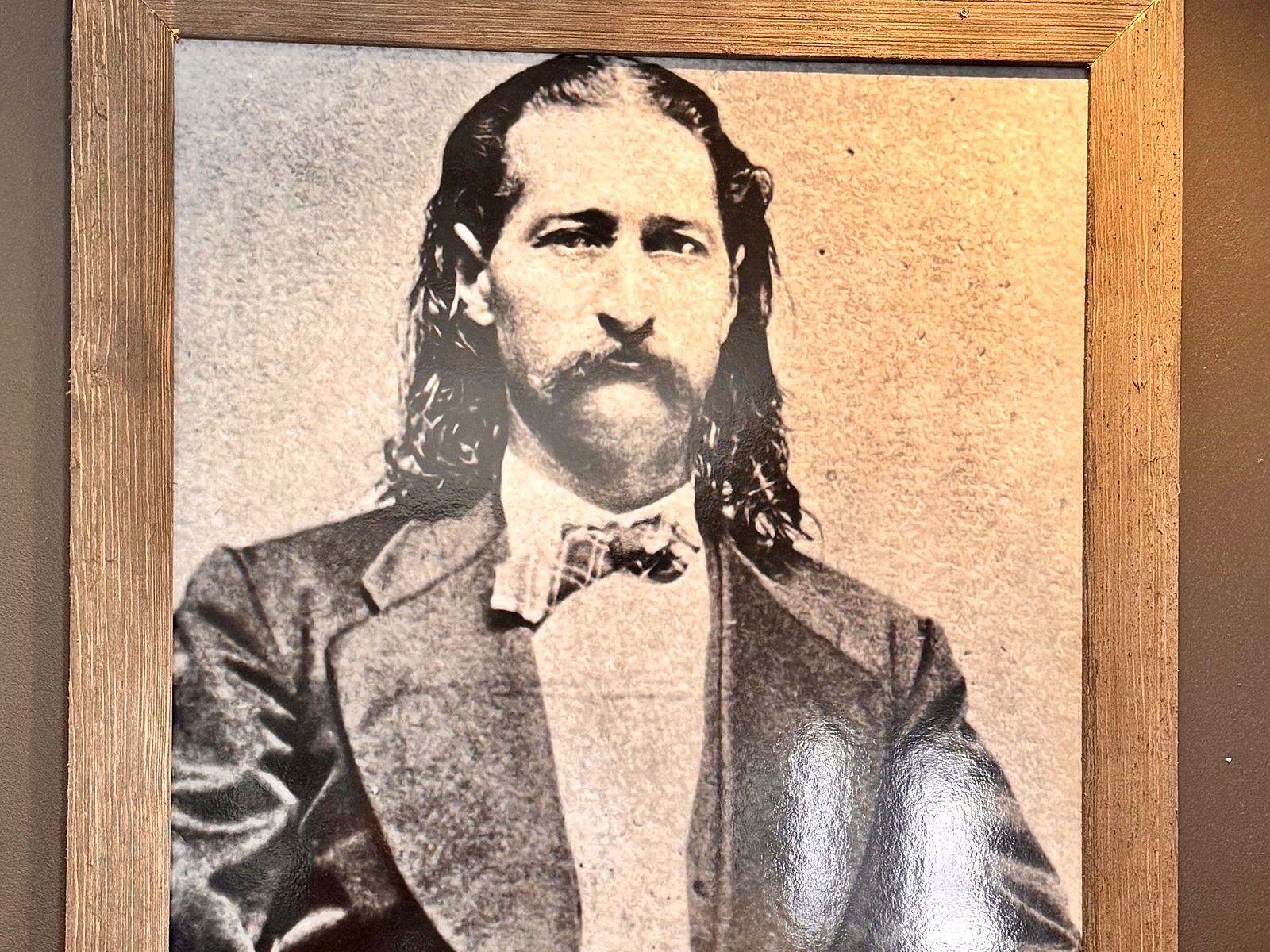 A framed portrait of Wild Bill Hickok on the wall at Hickok&rsquo;s — long hair, drooping moustache, frock coat and bow tie.