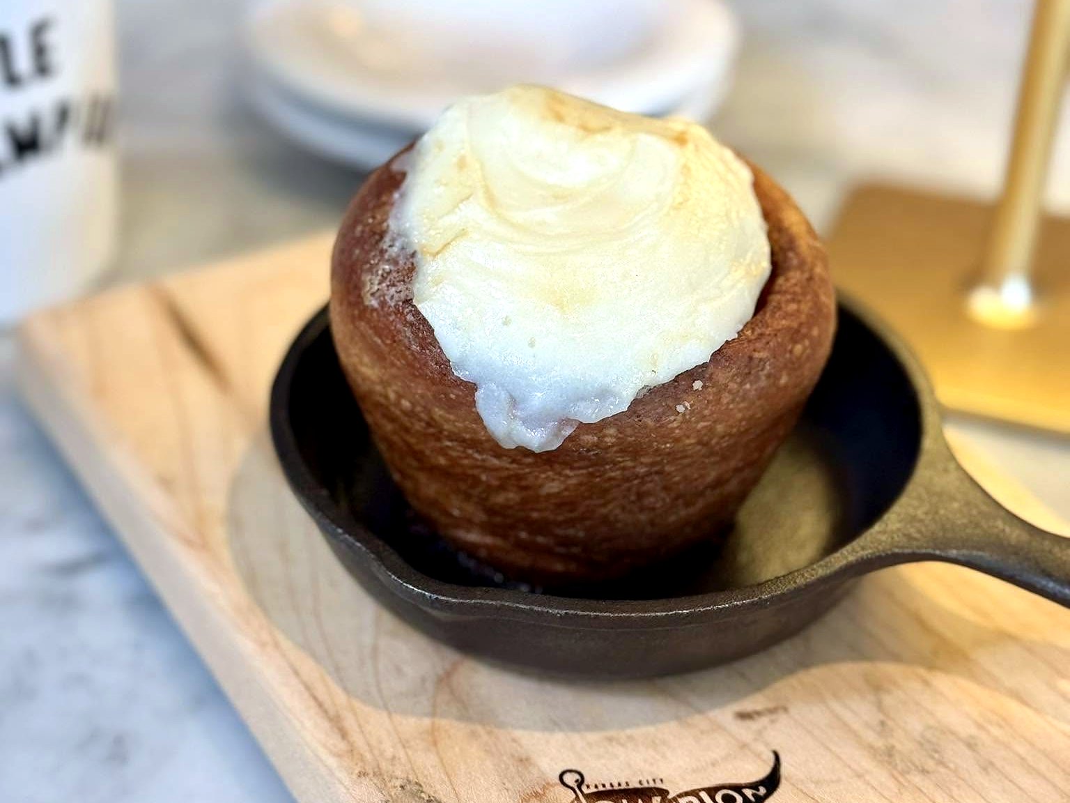 The cinnamon roll at Le Champion — a tall bronzed roll crowned with frosting, served in a mini cast-iron skillet on the café’s branded board.