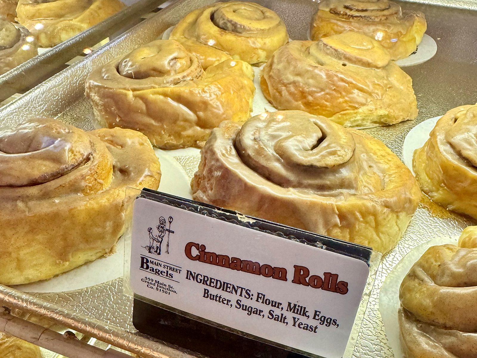 Cinnamon rolls at Main Street Bagels &mdash; thick glazed spirals, still on the bakery tray, beside a card listing the ingredients: flour, milk, eggs, butter, sugar, salt, yeast.
