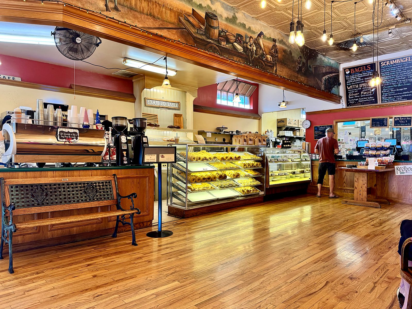 Inside Main Street Bagels — a pressed-tin ceiling, a painted mural running above the counter, pastry cases, and chalkboard menus over the espresso bar.