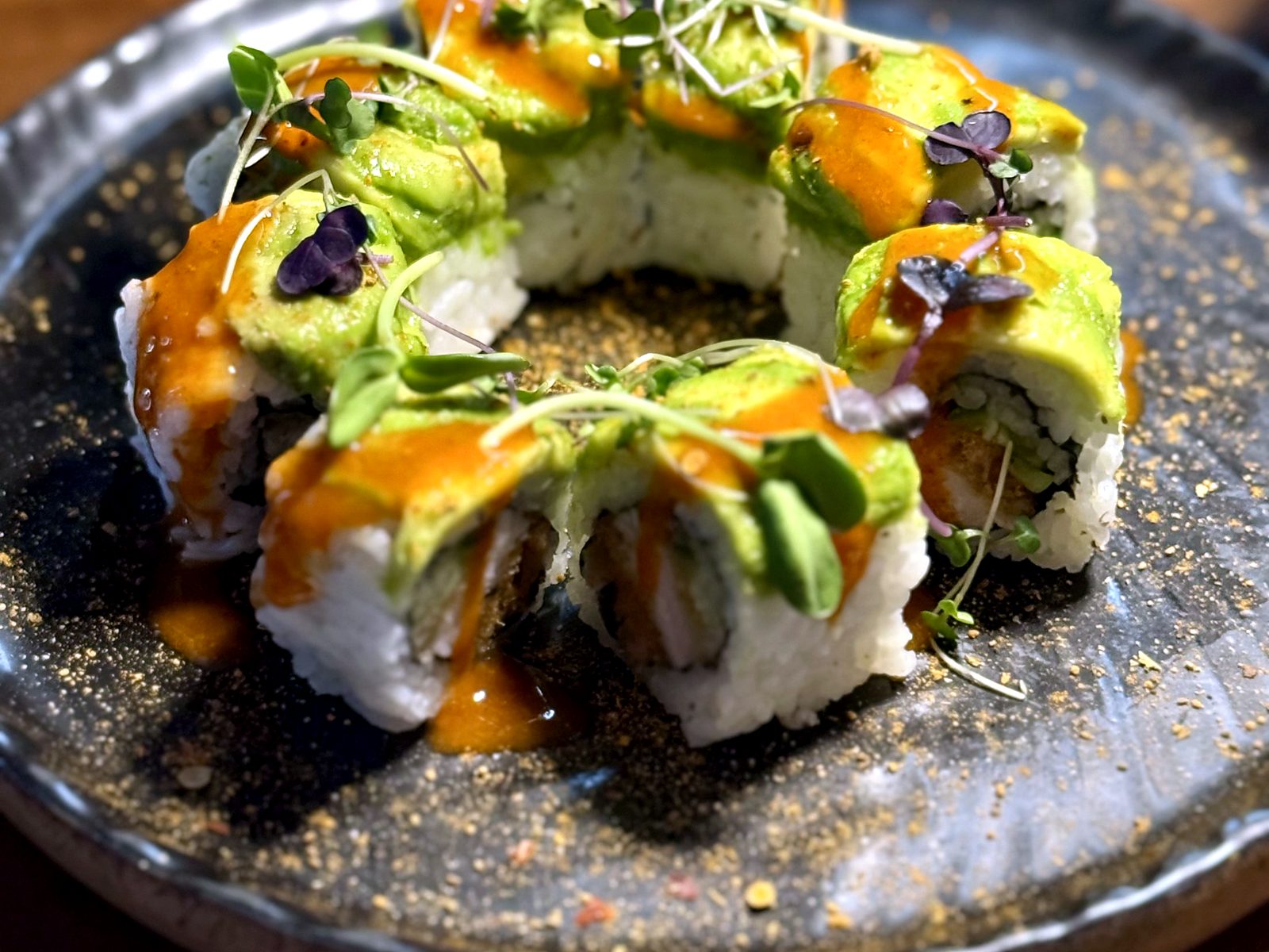 The alligator sushi roll at Noka — fried alligator inside, avocado draped over top, spicy sauce and microgreens, on speckled ceramic.