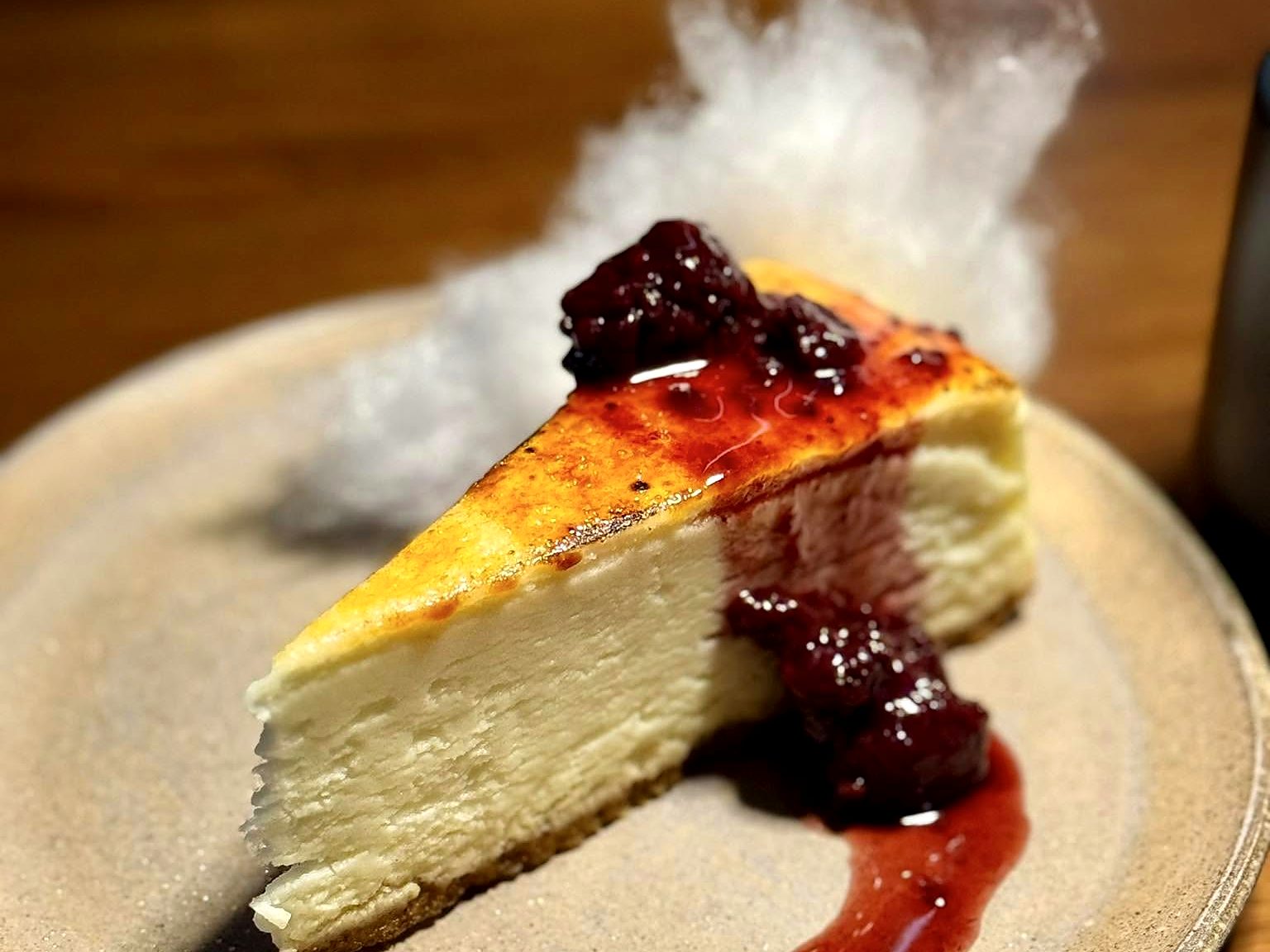 Cheesecake at Noka — a brûléed, burnt-top slice with dark berry compote, a wisp of spun sugar rising behind it.