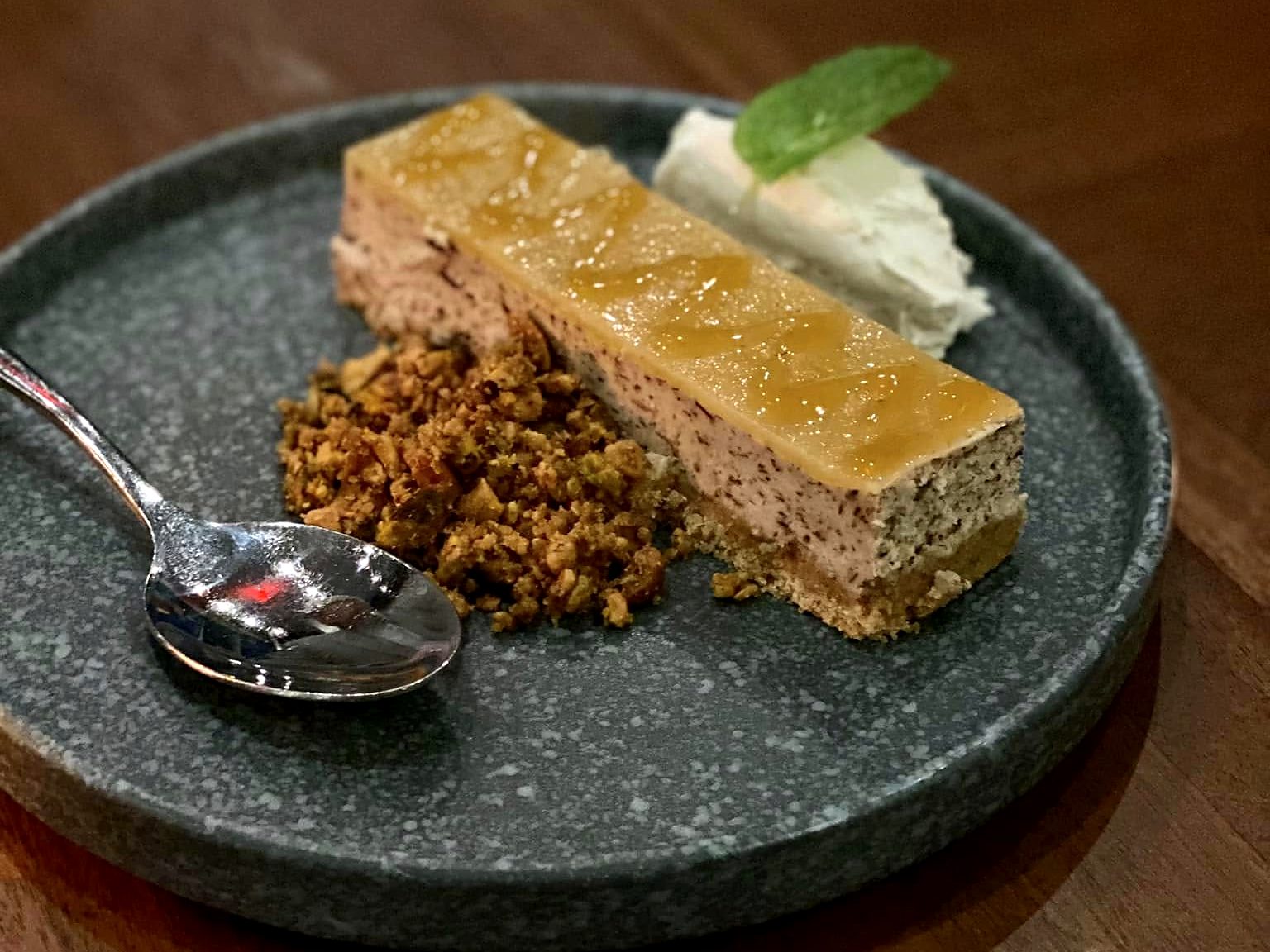 A glazed dessert bar at Of Course Kitchen & Company — spiced cake under a shining glaze with pistachio crumble, whipped cream, and mint.
