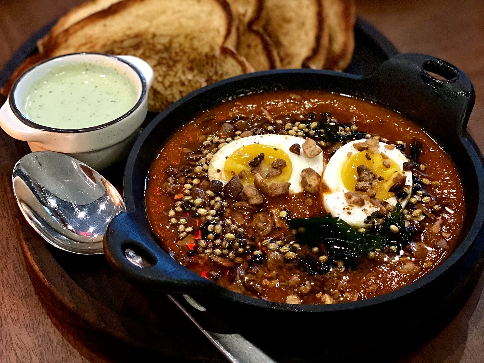 Egg curry toast at Of Course Kitchen & Company — jammy eggs in a cast-iron pan of deep red curry with toasted spices and curry leaves, toast fanned behind, herb cream alongside.