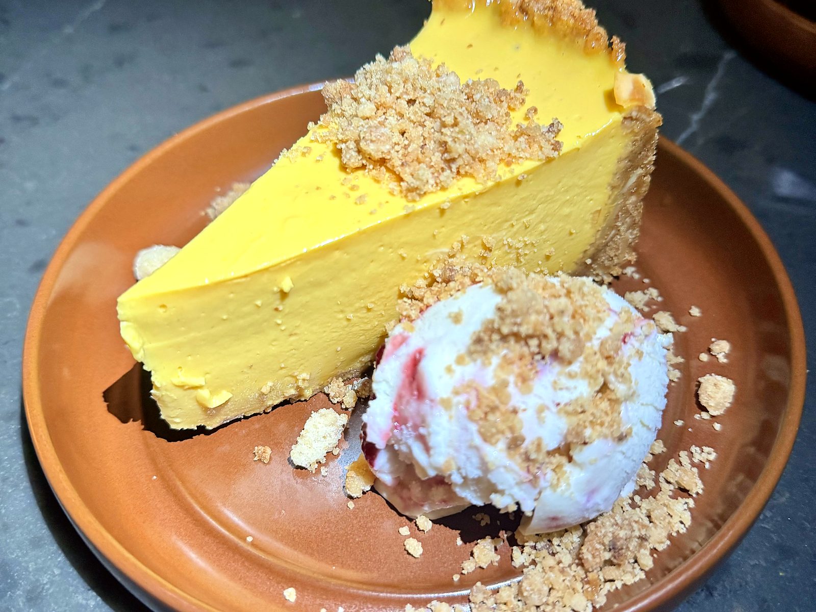 Mango pie at Of Course Kitchen & Company — a bright yellow slice in a graham crust under toasted crumble, beside a scoop of blackberry cheesecake ice cream.