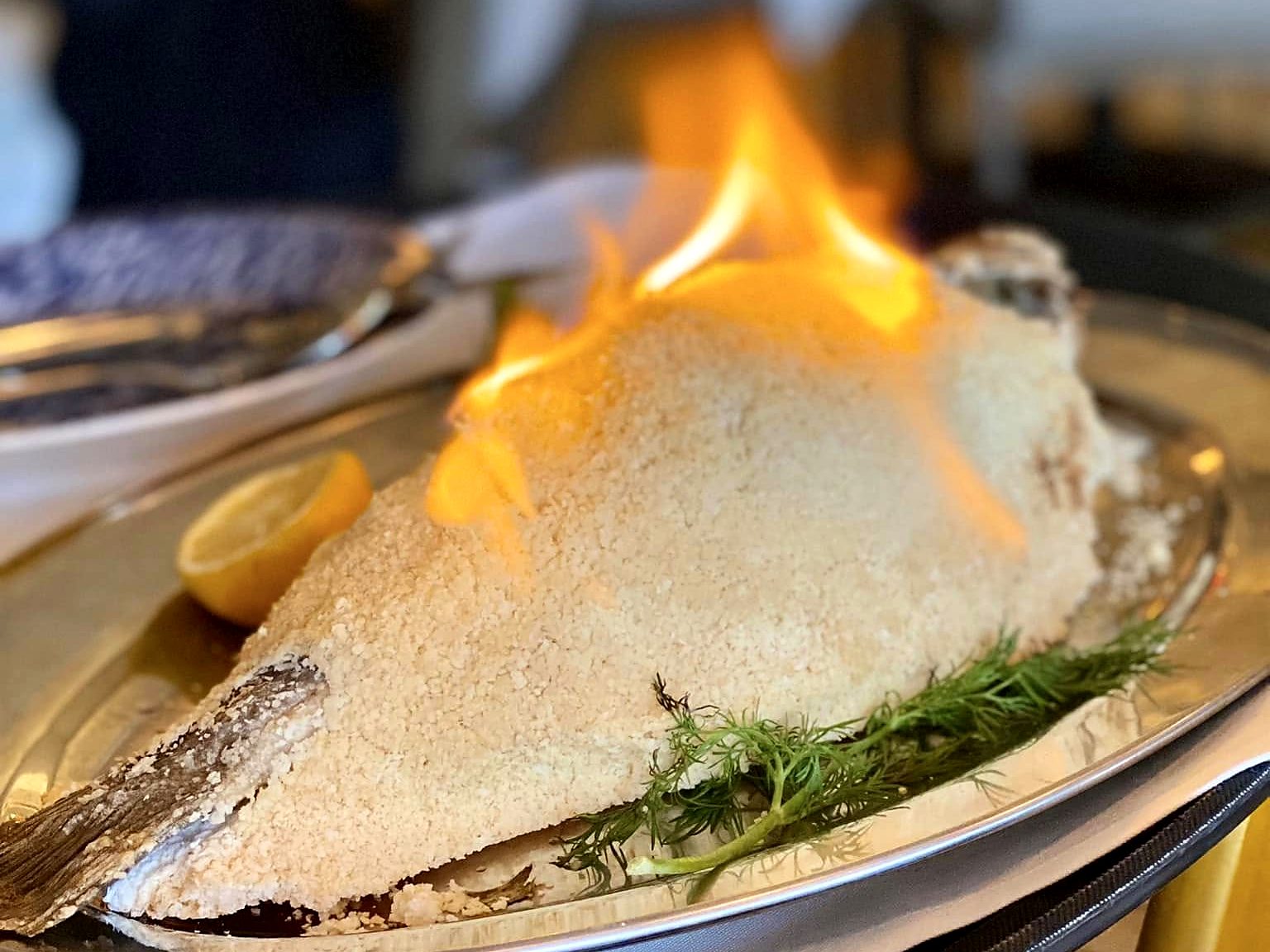 The sea-salt-crusted branzino at Paros, flambéed tableside — flames rising off the salt dome, dill and a lemon half alongside.