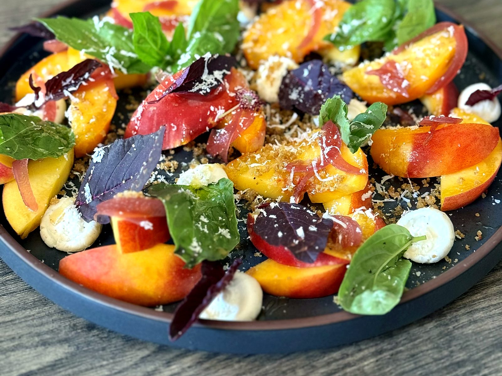 Peach caprese at Pêche — Palisade peaches with basil, purple shiso, goat cheese, cured ham, and shaved cheese on a dark plate.