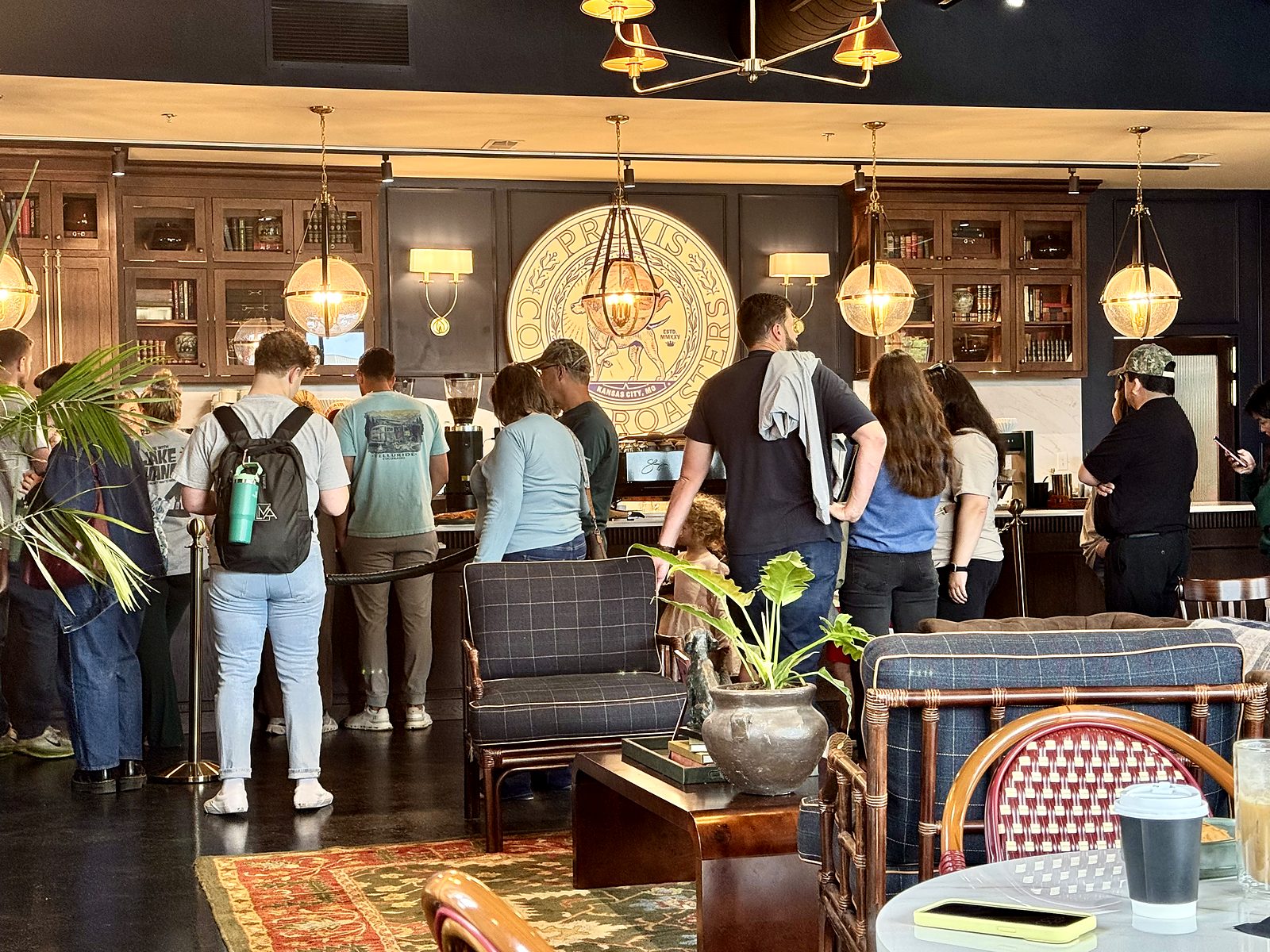 Inside Provis Coffee — a line at the counter under the round Provis Coffee Roasters seal, brass pendant lamps, glass-front cabinets of books, and plaid armchairs in the foreground.