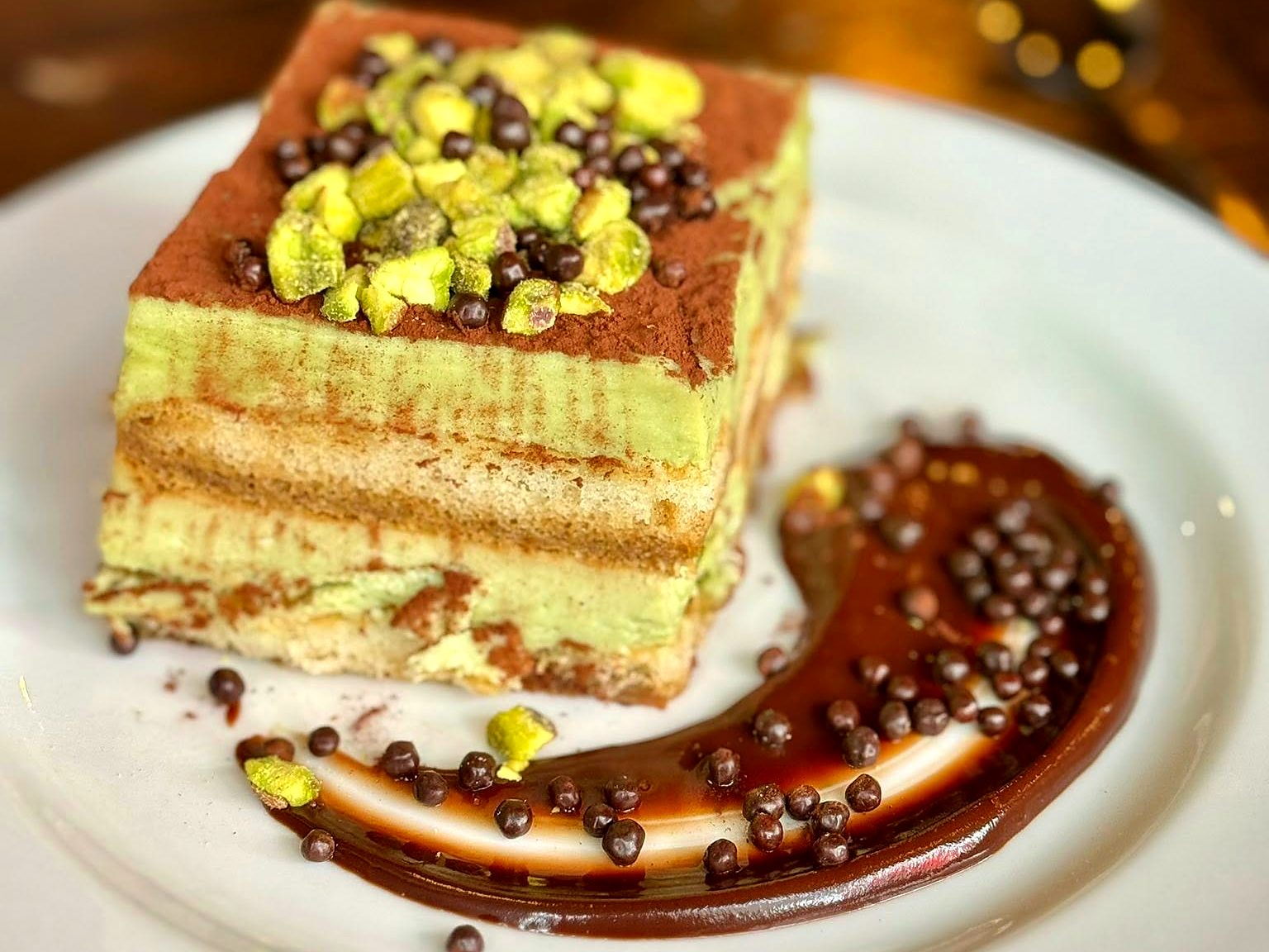 Pistachio tiramisu at The Pullman — cocoa-dusted layers of green pistachio cream and ladyfingers, crushed pistachios on top, a chocolate-pearl swoosh alongside.