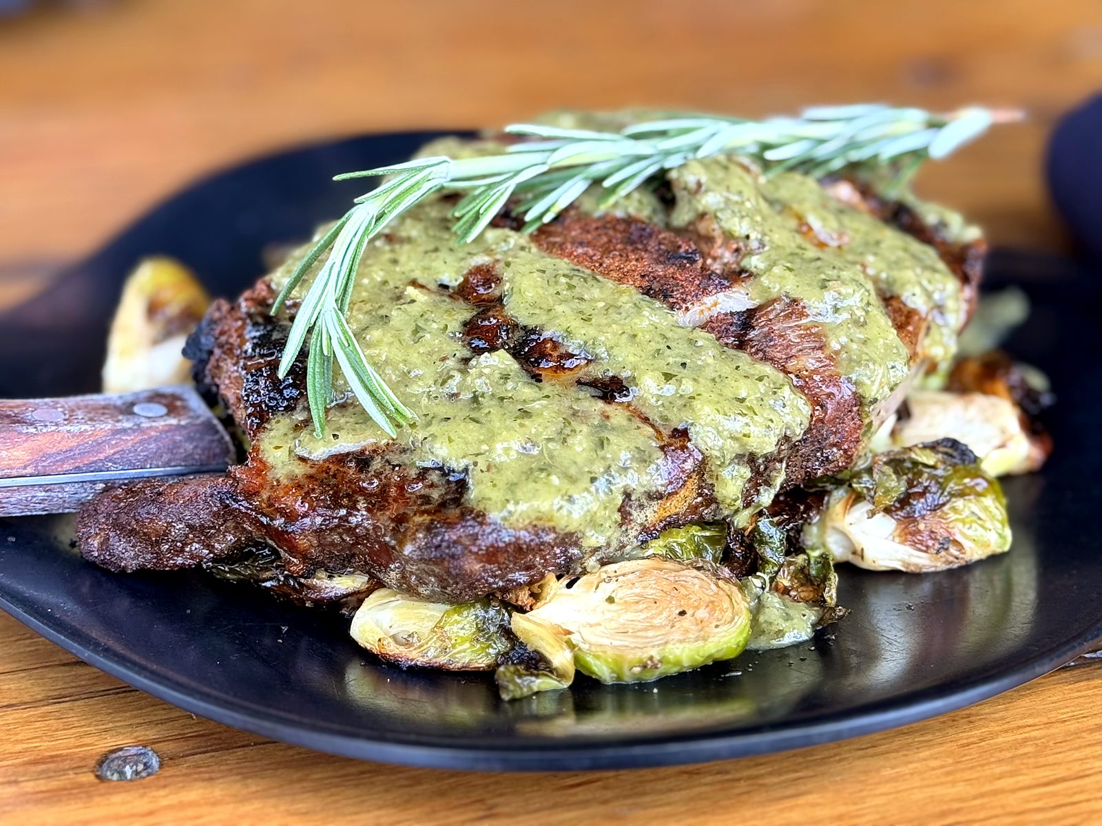A bone-in ribeye at The Russell draped in green salsa verde with rosemary, over roasted brussels sprouts on a black plate.