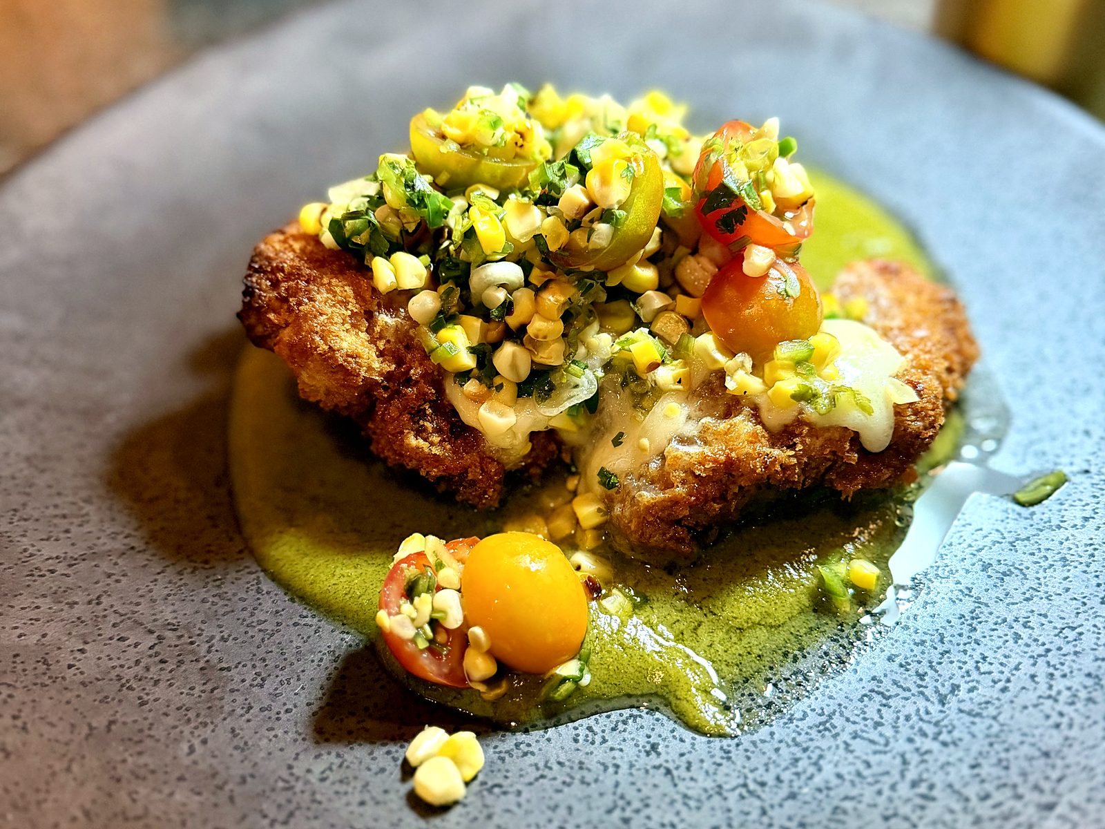The crispy pork cutlet at Scratch — panko-fried, under melted cheese, charred corn, and cherry tomatoes, set on a pool of green salsa.