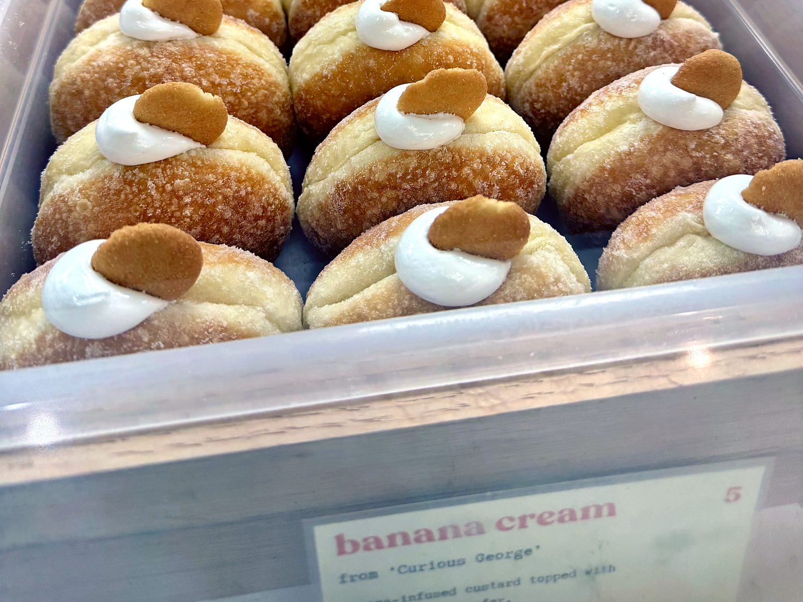 A tray of banana cream doughnuts at Slow Rise — sugar-tossed sourdough rounds, each topped with a swirl of cream and a vanilla wafer, above a flavor card crediting &lsquo;Curious George&rsquo;.