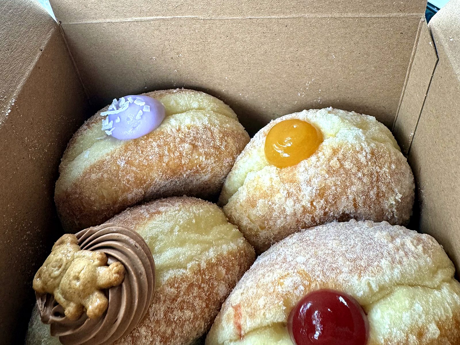 A box of four filled sourdough doughnuts from Slow Rise — one purple-frosted with coconut flakes, two glossy fruit-filled, and a chocolate swirl crowned with a tiny bear cookie.