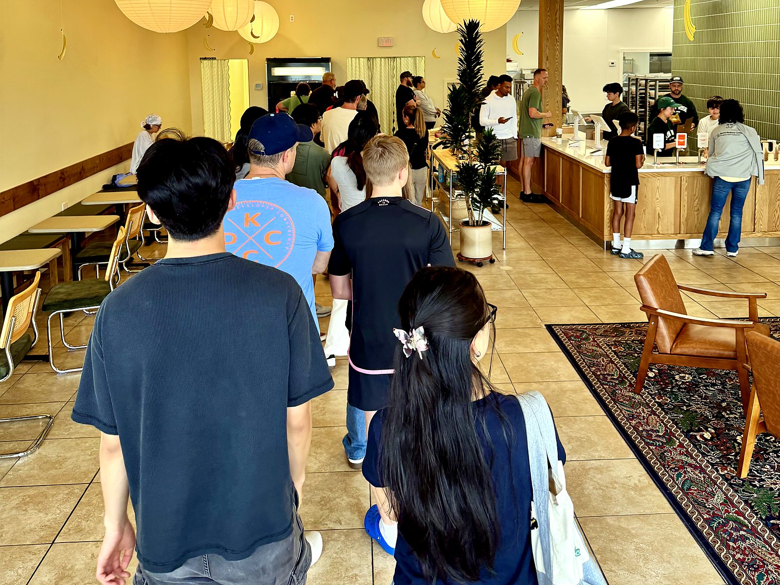 The morning line inside Slow Rise in Overland Park — customers queued past the tables under paper lanterns and hanging banana decorations while staff box doughnuts at the counter.