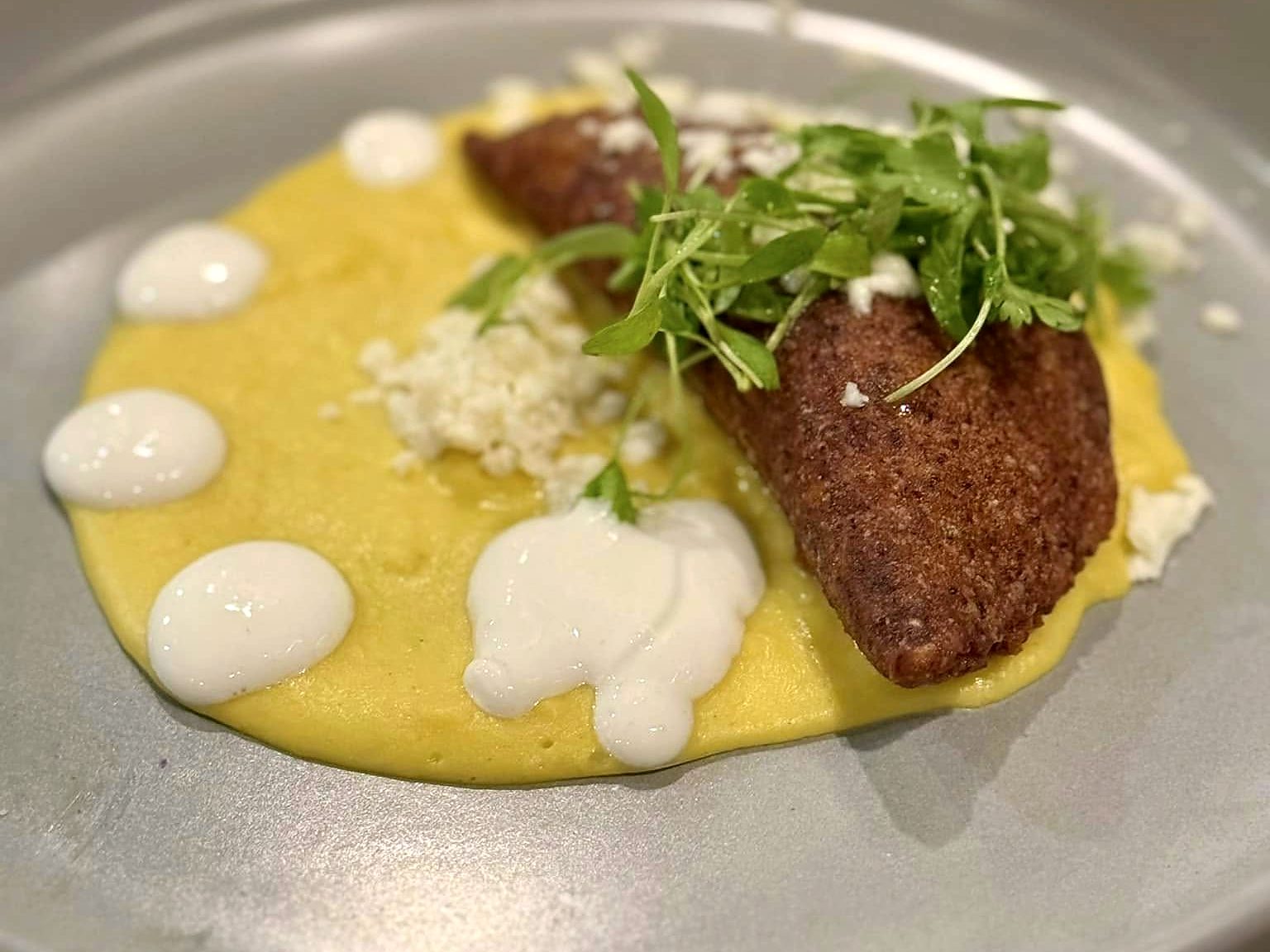 An empanada at T’ähä — crisp-fried and golden on a sweep of yellow purée dotted with crema, scattered with queso fresco and cilantro sprigs.