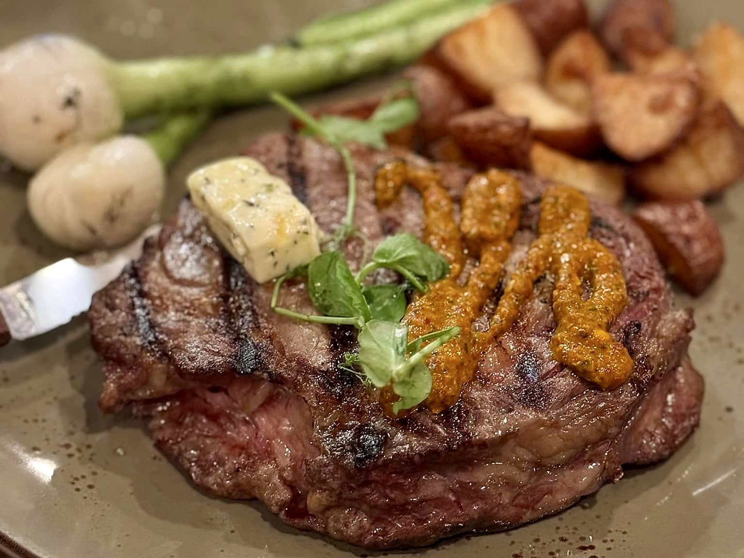 A grilled ribeye at T’ähä, striped from the fire and finished with herb butter and a ribbon of adobo, with roasted potatoes, spring onions, and asparagus.