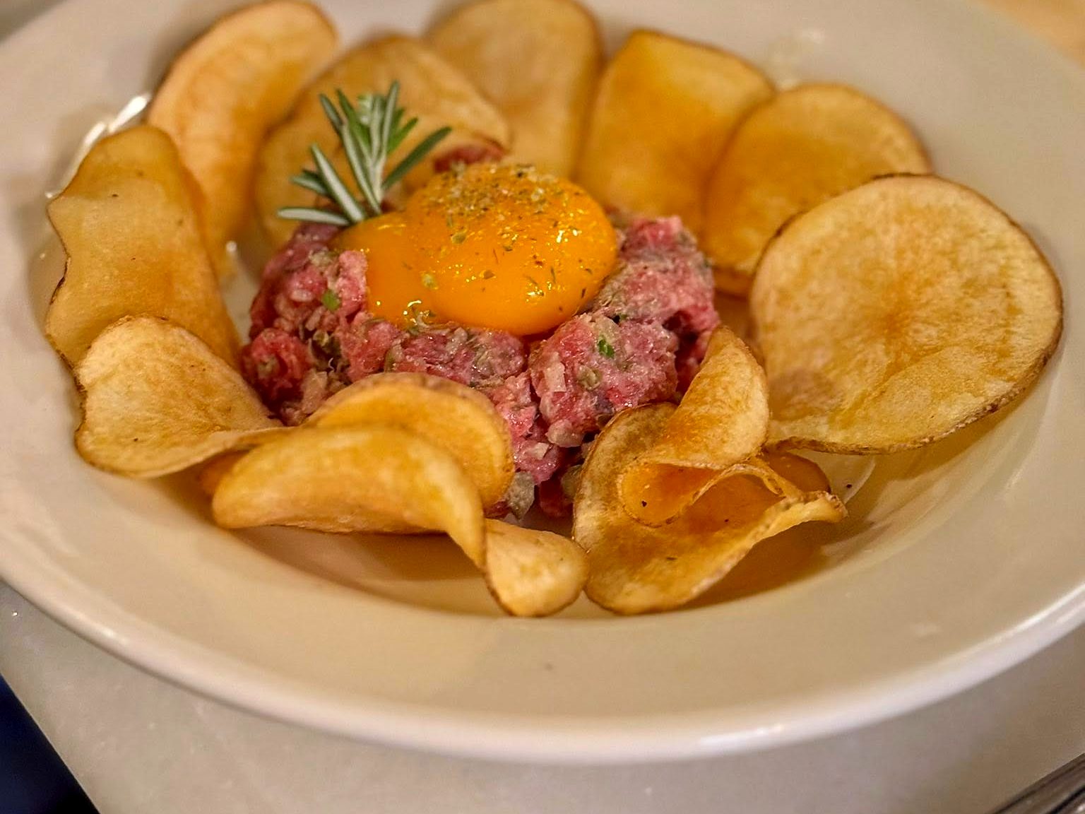 Steak tartare at Tailleur — a sunset-orange yolk over hand-chopped beef and rosemary, ringed by house potato chips.