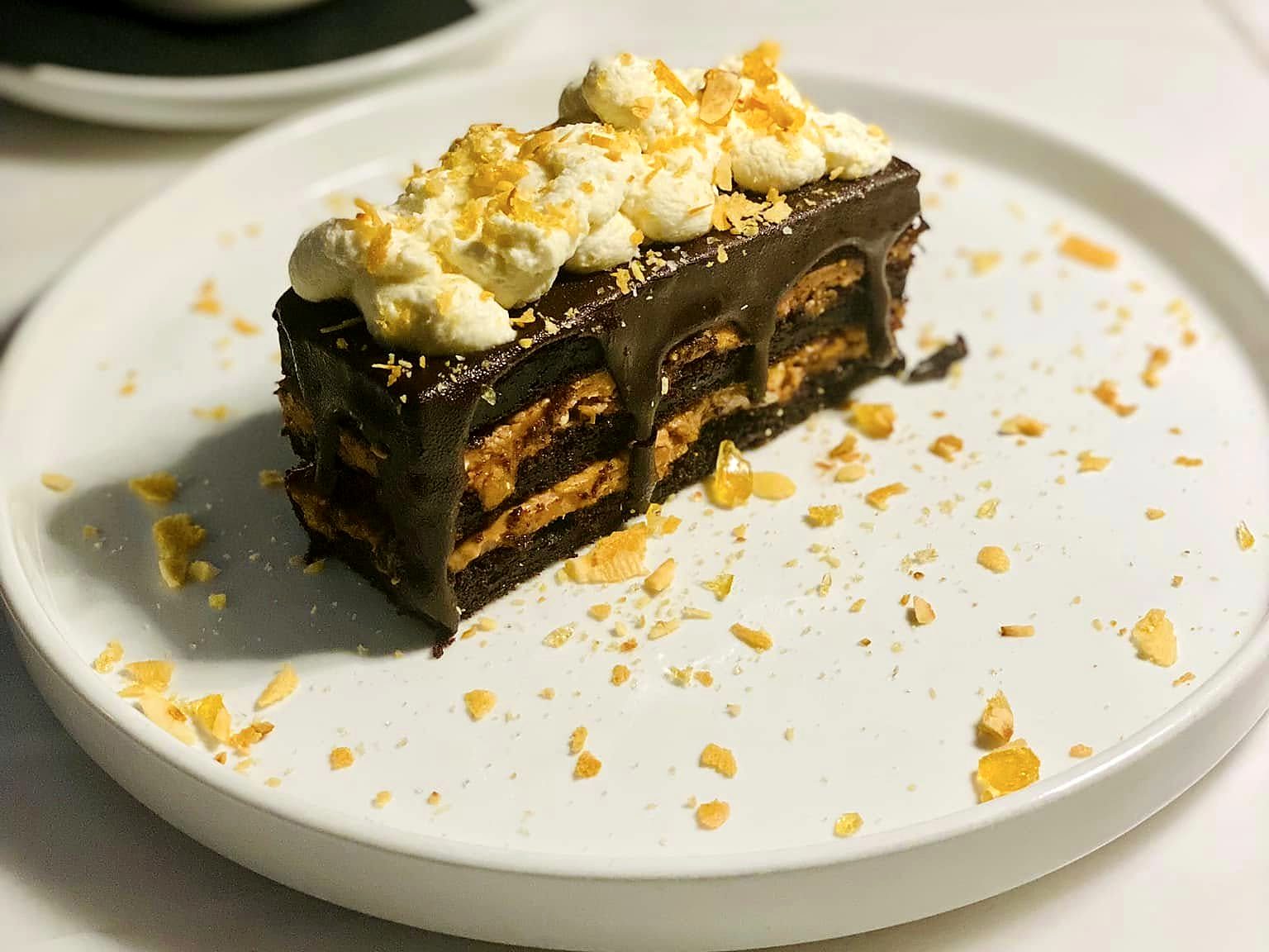 A chocolate layer cake at Verbena — ganache-glazed with peanut butter layers, whipped cream and toffee shards on top, toffee crumbs scattered alongside.