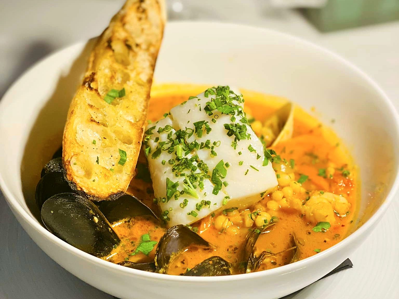 Cioppino at Verbena — a white fish fillet under chives and parsley in saffron broth with mussels, clams, and pearl couscous, with grilled baguette alongside.